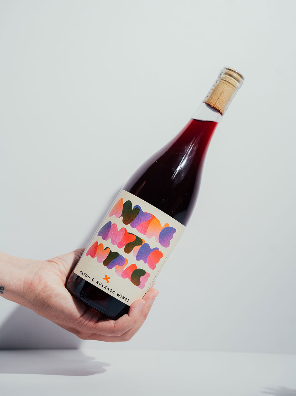 Bottle of Catch & Release Wine Anyone Anytime Anyplace, red blend, with a colorful label held by a hand on a light gray background