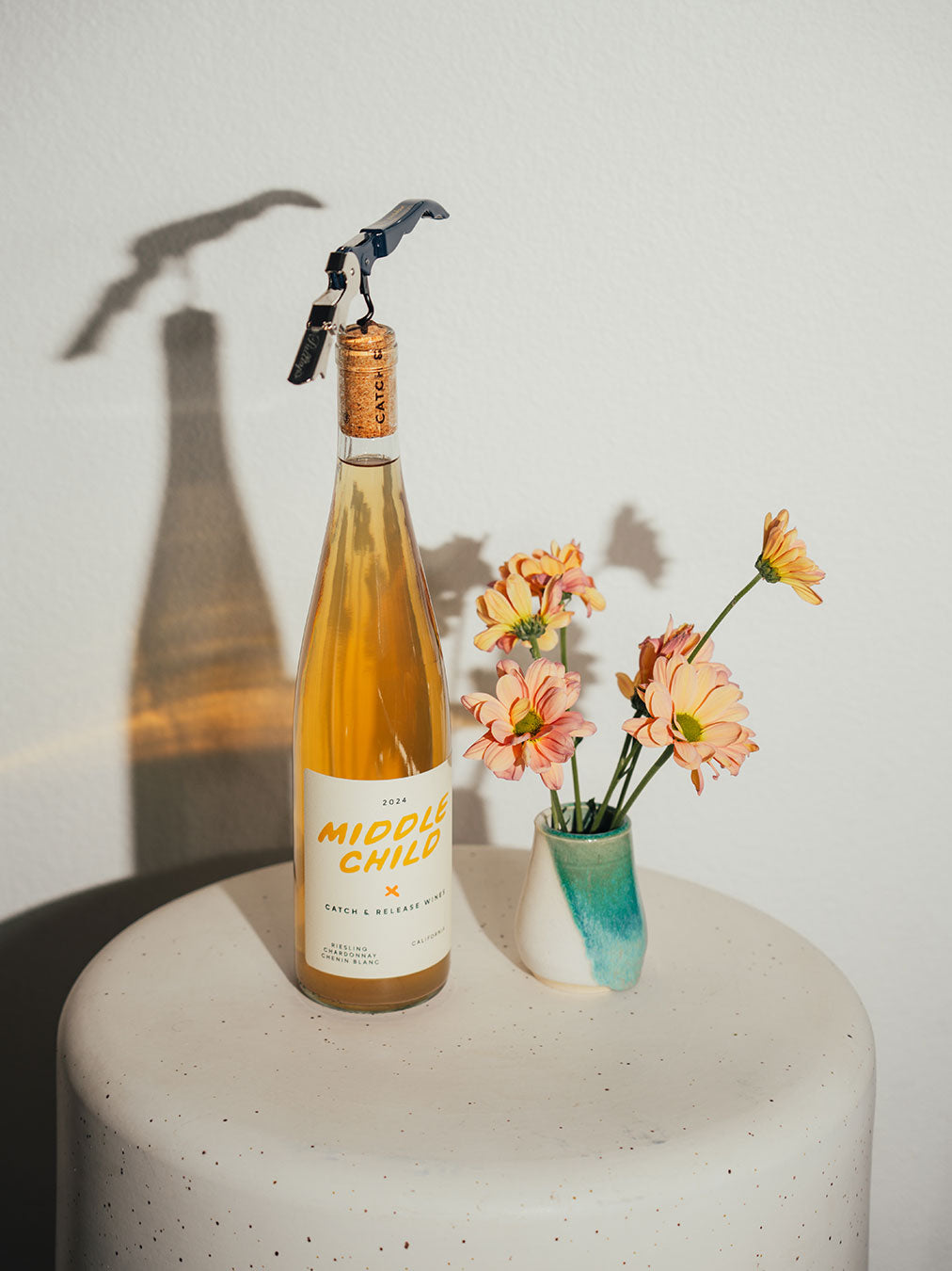 Bottle of Catch & Release Wines Middle Child orange wine with a cork on a white surface next to a small vase with flowers.