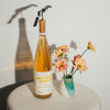 Bottle of Catch & Release Wines Middle Child orange wine with a cork on a white surface next to a small vase with flowers.